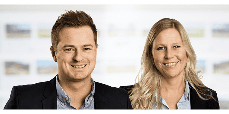 Byliv.nu: To personer, en mand og en kvinde, står side om side, begge smilende. De er klædt i forretningstøj og fremstår i et godt oplyst indendørs miljø med slørede baggrundsbilleder.