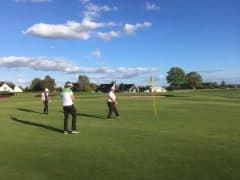 Byliv.nu: Tre golfspillere på en green, nær en flagstang, under en klar blå himmel med spredte skyer. Huse og træer er synlige i baggrunden.