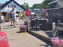 Byliv.nu: Folk sidder og står uden for en fiskerestaurant med skilte, hvor der står "FRISK." Der er blomster i plantekasser og parkerede biler i baggrunden.