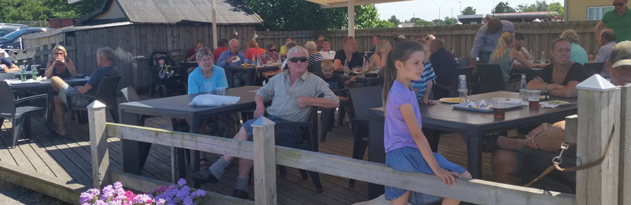 Byliv.nu: Folk sad ved udendørs borde på en restaurant på trædæk, socialt og nyder mad under en hvid baldakin. Blomster i krukker står langs trærækværket i forgrunden.