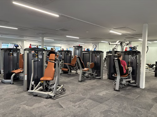 Byliv.nu: Folk, der bruger træningsmaskiner i et fitnesscenter med nummereret udstyr og overheadbelysning.